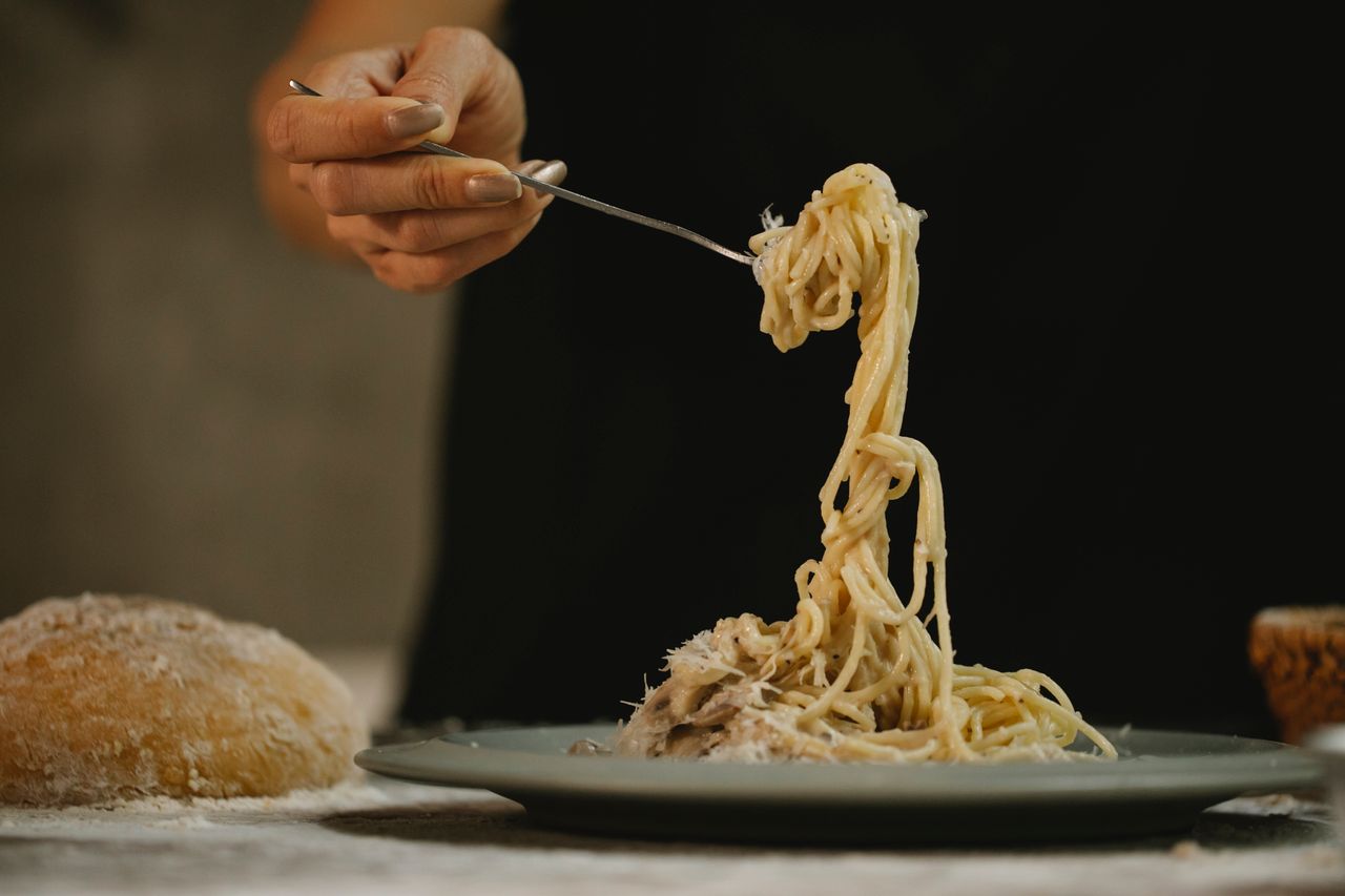 Crop anonymous female with fork enjoying tasty spaghetti with cream mushroom sauce served on table near freshly baked bread